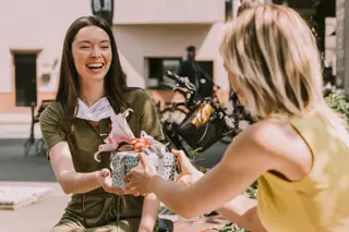 Laughing woman with taken off face mask handing over gift to friend